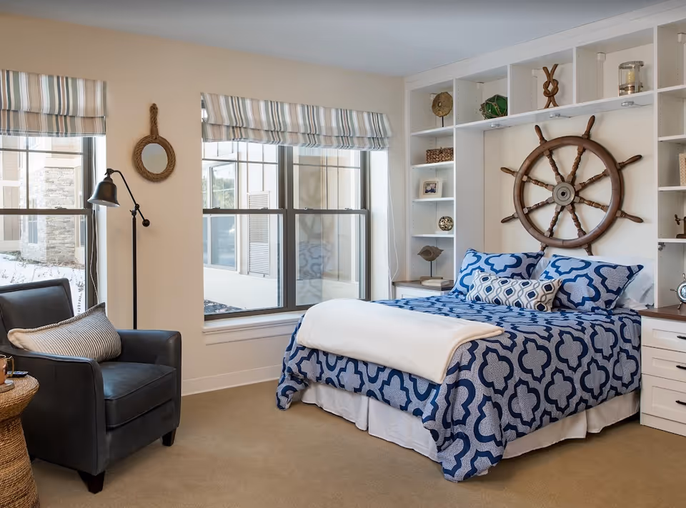 Bright nautical-themed bedroom featuring a blue patterned bed, built-in white shelving with a ship's wheel above the headboard, a window, and a leather armchair.