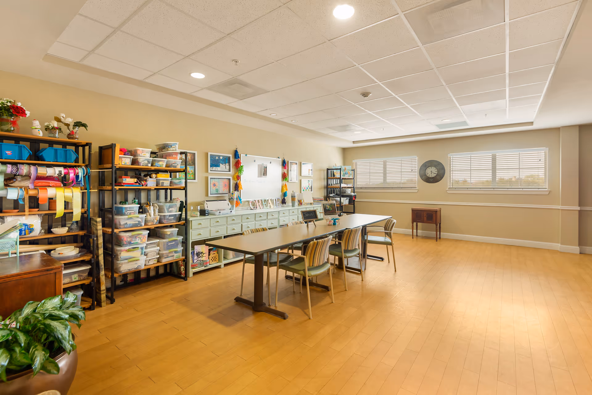 A bright and spacious activity room with wooden flooring and beige walls. There are two tables pushed together with six chairs around them. On the left side, there are shelves filled with craft supplies including ribbons, containers, and various art materials. The back wall features a whiteboard and framed artwork. Two large windows with blinds let in natural light, and a clock is mounted on the wall between the windows. A small wooden cabinet is positioned under the windows.
