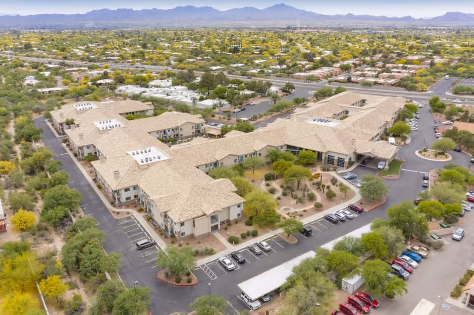 Aerial view of Mountain View Retirement Village showing a large, beige-roofed building complex surrounded by parking lots, trees, and landscaped areas with mountains visible in the background under a clear sky.