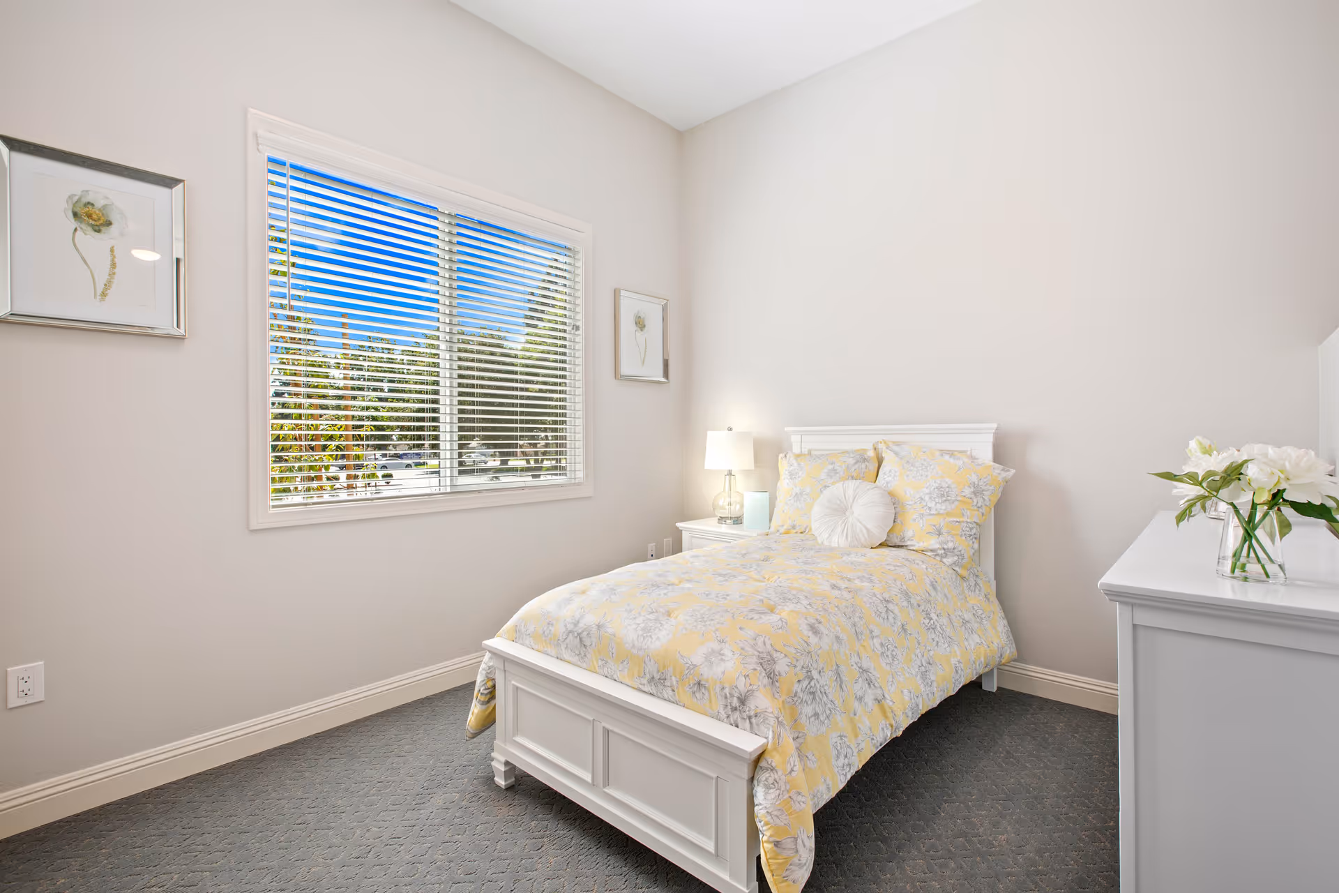 A bright bedroom with a single white bed featuring yellow and gray floral bedding. There is a white nightstand with a lamp and a small book, a white dresser with a vase of white flowers, and two framed floral artworks on the light beige walls. A large window with white blinds shows a view of trees and a blue sky outside.