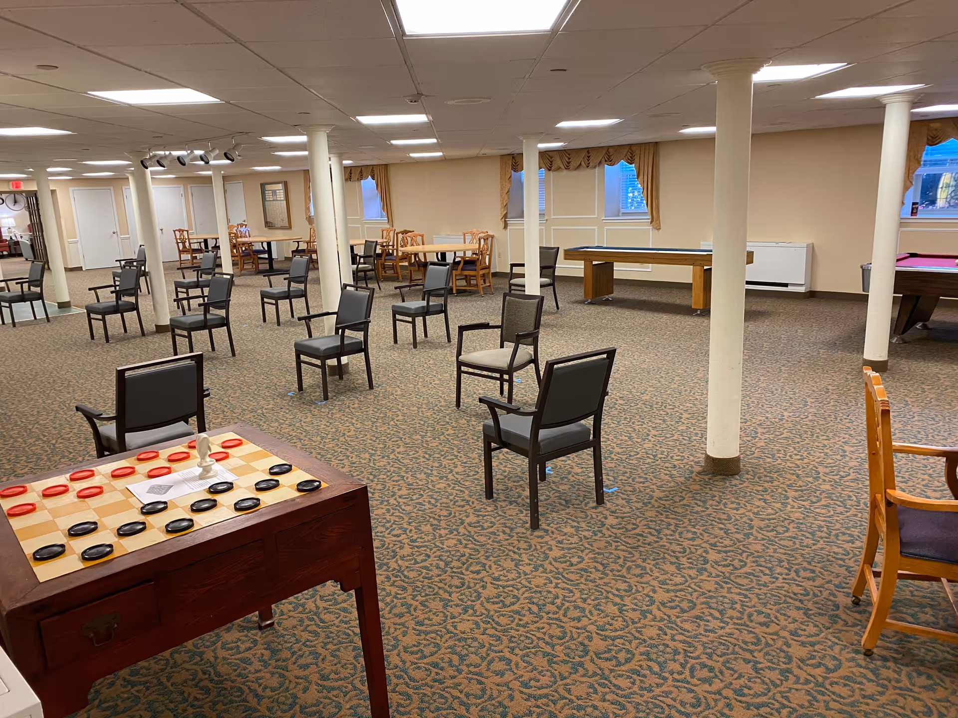 A spacious common room with multiple chairs arranged in small groups and around tables. There is a large checkers board on a wooden table in the foreground, and in the background, there are additional tables and chairs, as well as a pool table and a shuffleboard table. The room has beige walls, patterned carpet, and several white support columns.
