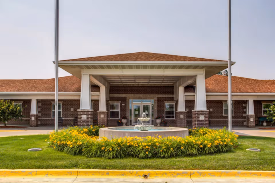 Front exterior view of Prairie Hills at Independence facility showing a brick building with a covered entrance, two flagpoles, a circular flower bed with yellow flowers, and a water fountain in the center.