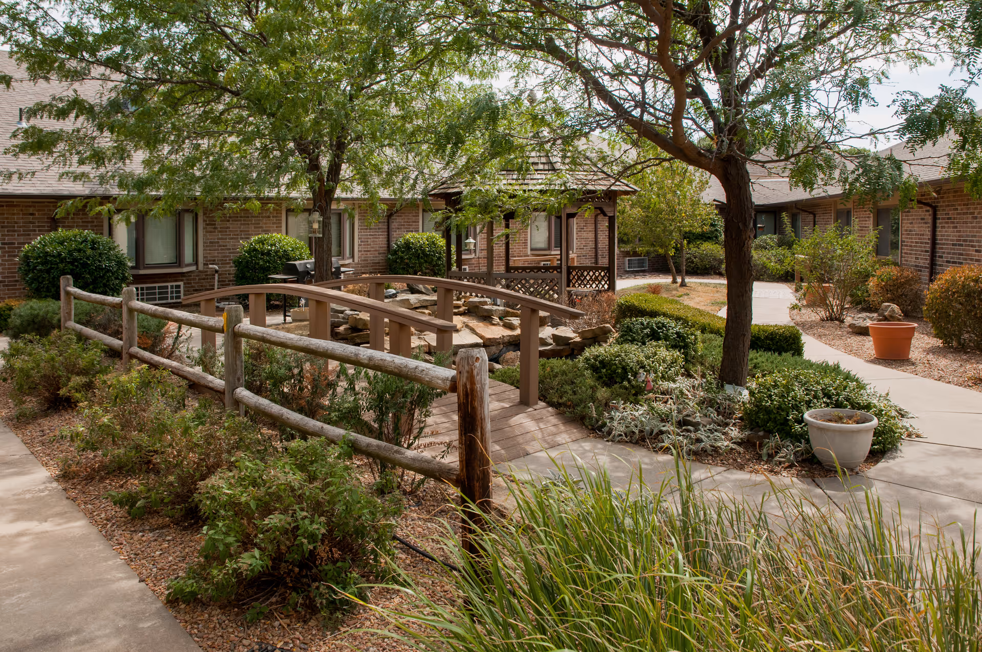 A peaceful outdoor courtyard area with a small wooden bridge over a dry rock bed, surrounded by shrubs, trees, and potted plants. Brick buildings with windows and a covered patio are visible in the background.
