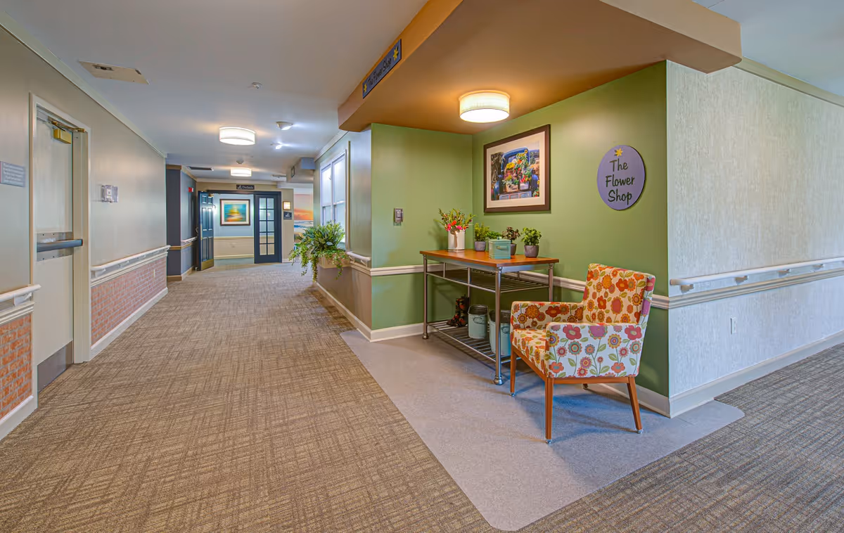 A well-lit senior living facility hallway with beige and green walls, carpeted floors, and handrails along the walls. On the right side, there is a small seating area with a colorful floral armchair and a metal table holding potted plants and flowers. A round sign on the green wall reads 'The Flower Shop'. The hallway extends into the distance with doors and framed artwork visible.