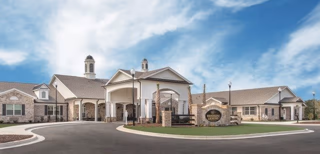 Front exterior view of Benton House of Aiken, a single-story senior living facility with a covered entrance, brick and siding exterior, and a circular driveway under a blue sky with some clouds.