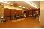 Interior view of a spacious room with wooden flooring, ceiling fans, large windows with blinds, and exercise equipment including parallel bars and a treadmill, suggesting a rehabilitation or therapy area.