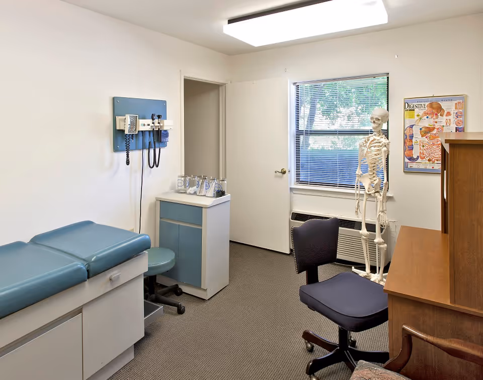 A medical examination room with a blue padded exam table on the left, medical instruments mounted on the wall, a small cabinet with glass jars, a black office chair, a wooden desk, a human skeleton model, and a digestive system poster on the wall near a window with blinds.
