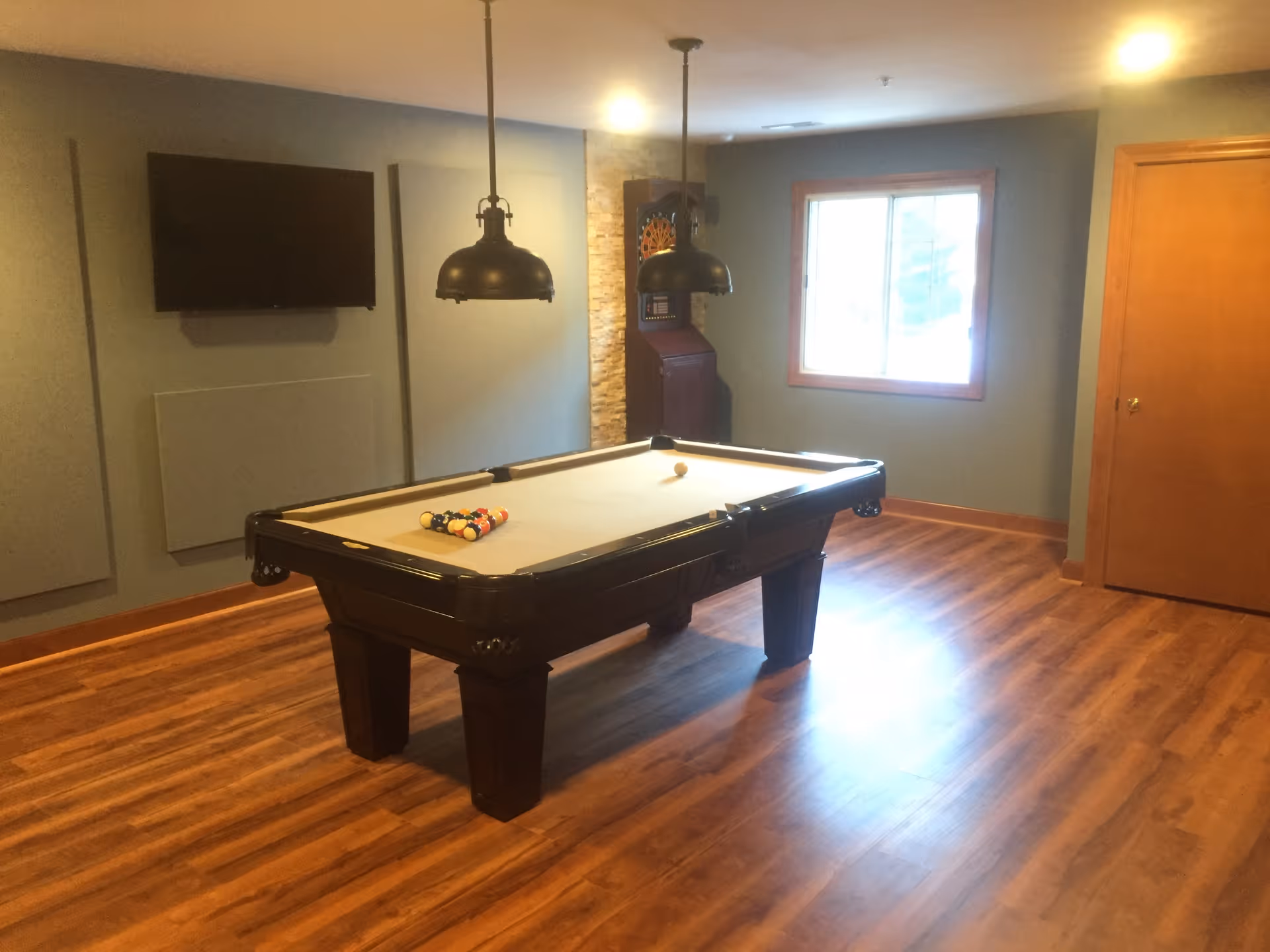 A recreation room with a pool table set up for a game, two hanging pendant lights above the table, a wall-mounted TV, a dartboard in the corner, a window letting in natural light, and wooden flooring.