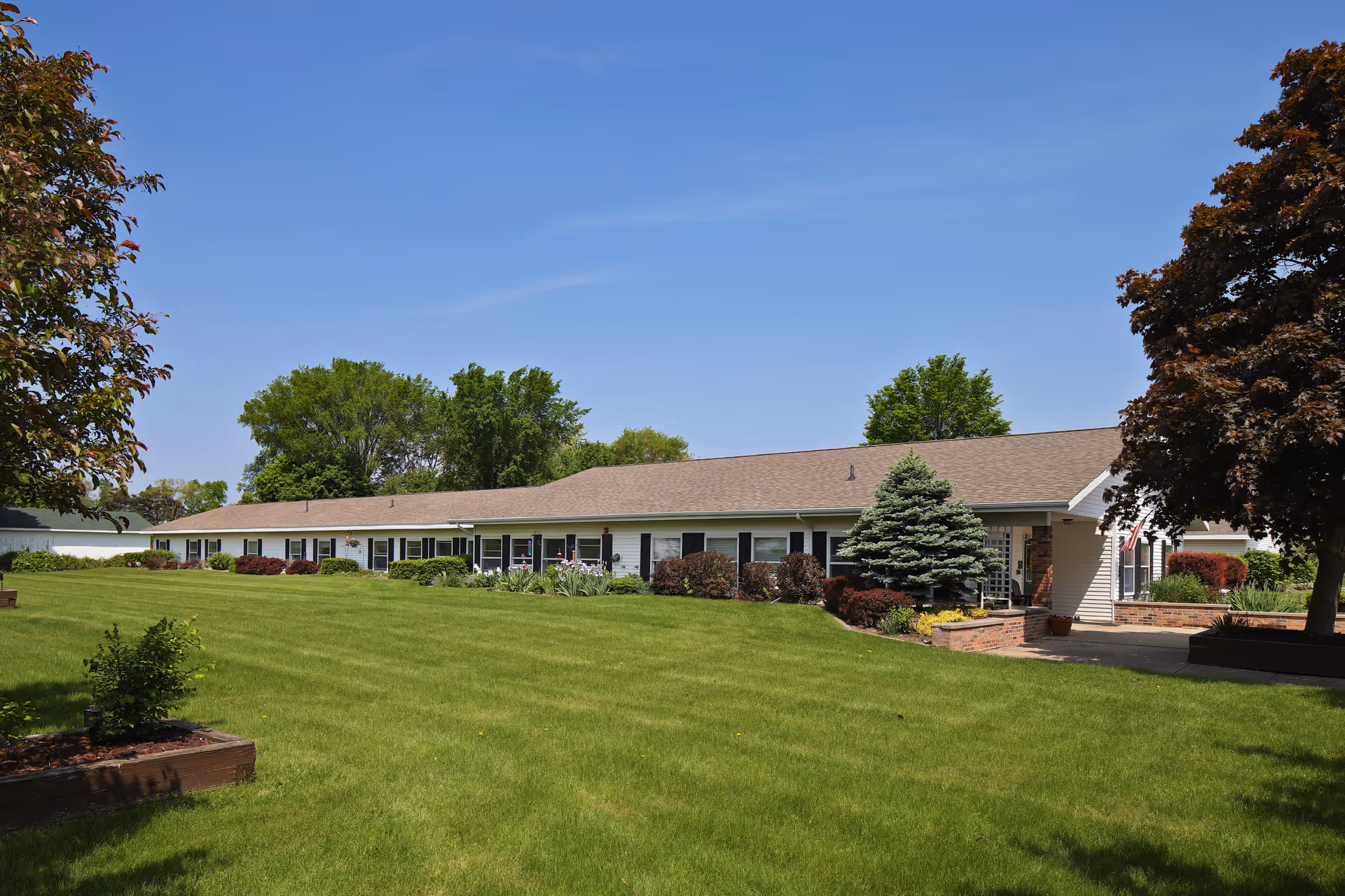 Single-story building with a long row of windows and a sloped roof, surrounded by well-maintained green lawn, shrubs, and trees under a clear blue sky.