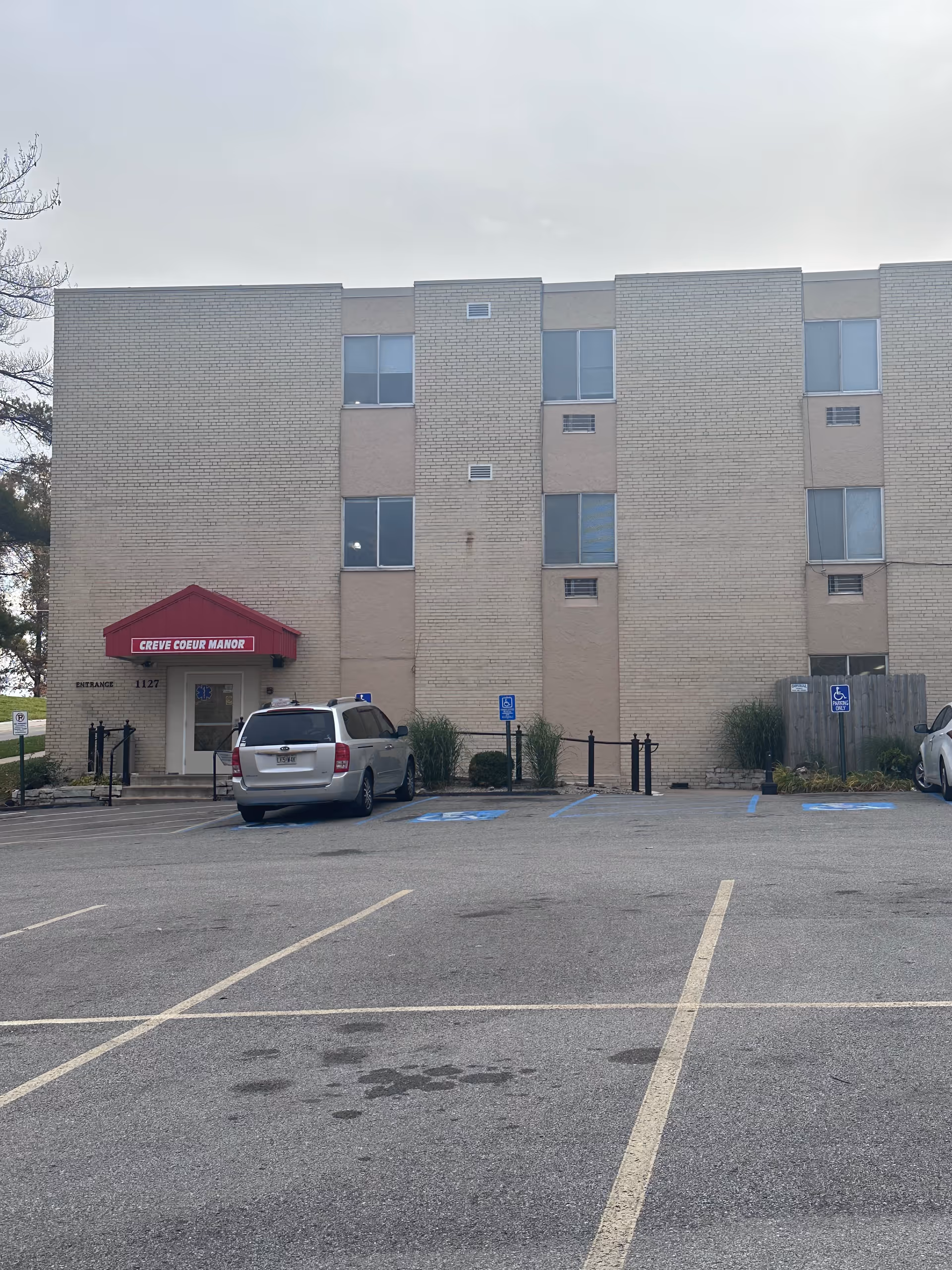 Exterior view of a three-story beige brick building with a red awning above the entrance that reads 'Creve Coeur Manor'. There are several windows and a parking lot in front with a few cars parked, including handicap parking spaces.