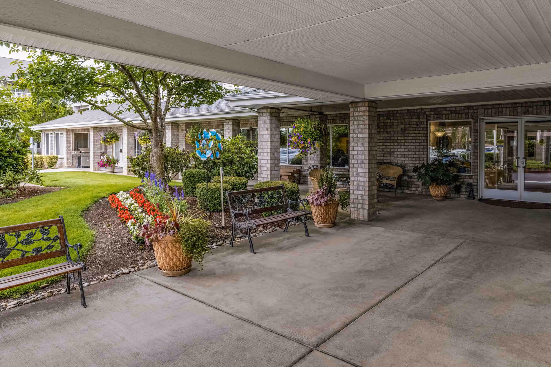 Covered entrance area of a senior living facility with benches, potted plants, hanging flower baskets, and a landscaped garden with colorful flowers and a tree. The building has brick walls and large windows, with double glass doors leading inside.