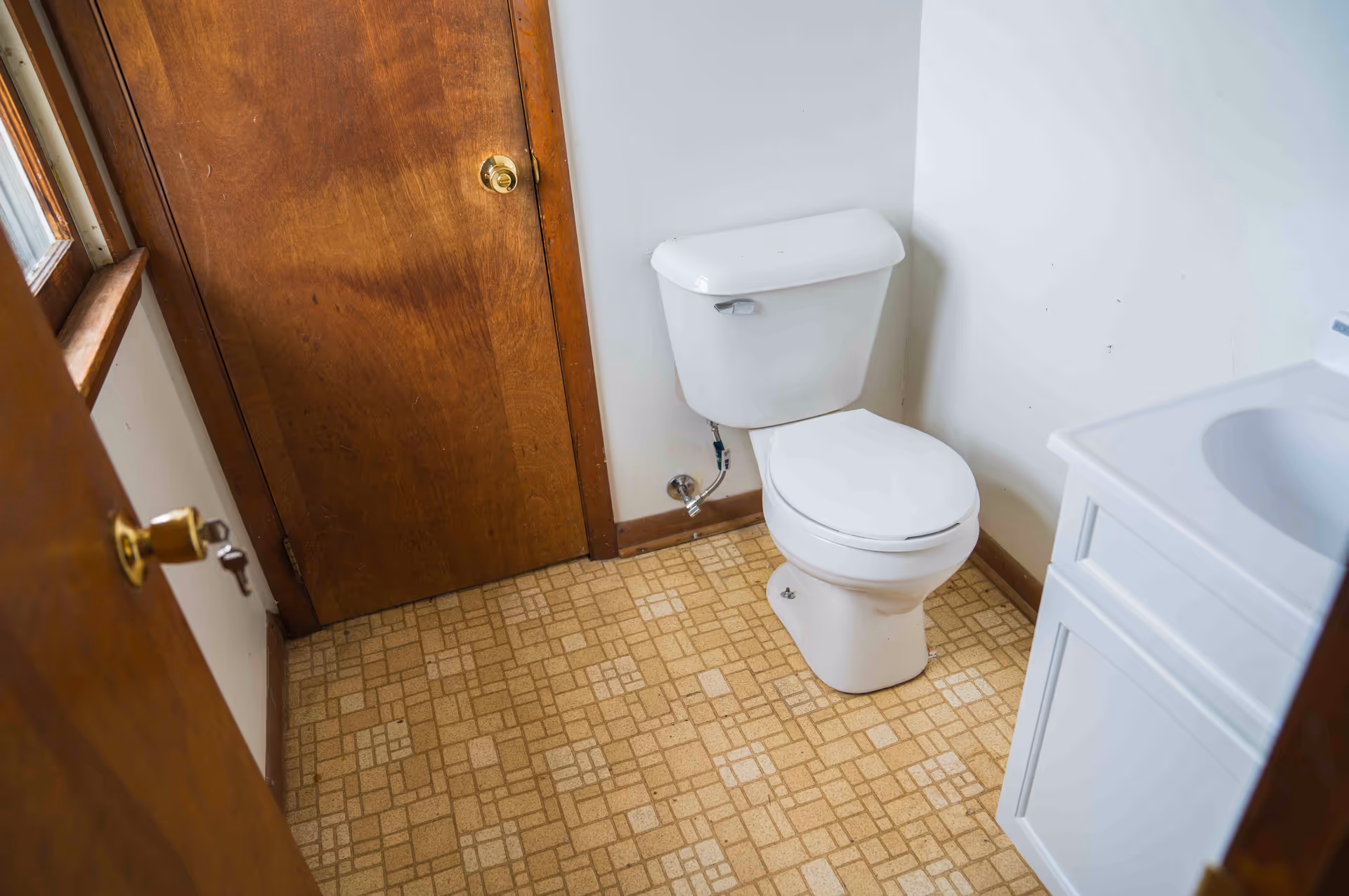 Small bathroom with a white toilet, a white sink cabinet, a wooden door, and a window. The floor has a patterned linoleum covering.