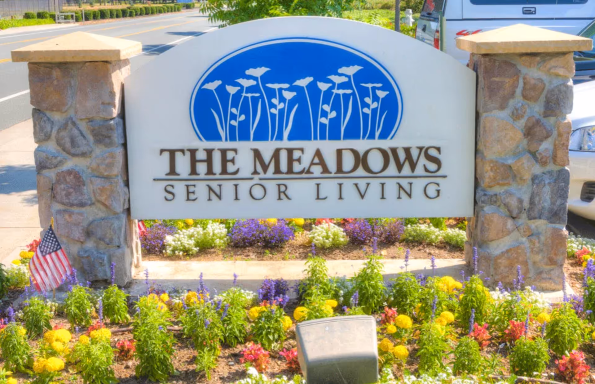 Stone pillars supporting a sign for The Meadows Senior Living, with colorful flowers planted in front and a small American flag on the left side. A road and some vehicles are visible in the background.