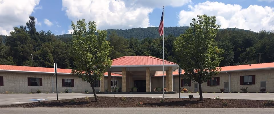 Front exterior view of Dogwood Crossing Senior Living facility with a single-story building featuring a red roof, beige brick walls, several windows with red shutters, an entrance with a covered portico, an American flag on a flagpole, and trees in front. Hills and a partly cloudy sky are visible in the background.