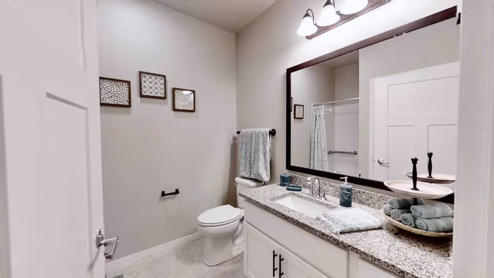 A clean and modern bathroom featuring a white toilet, a granite countertop with a sink, a large mirror with dark frame, and a three-light fixture above. The countertop holds neatly folded towels, soap dispensers, and a tiered tray with additional rolled towels. The walls are light-colored with three small framed artworks, and a towel hangs on a rack beside the toilet. A shower with a curtain is visible in the reflection of the mirror.