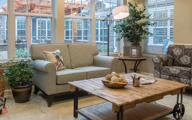 A cozy living room area with a beige loveseat adorned with a bird-patterned pillow, a patterned armchair, a wooden coffee table with decorative items, a floor lamp, and potted plants near large windows letting in natural light.