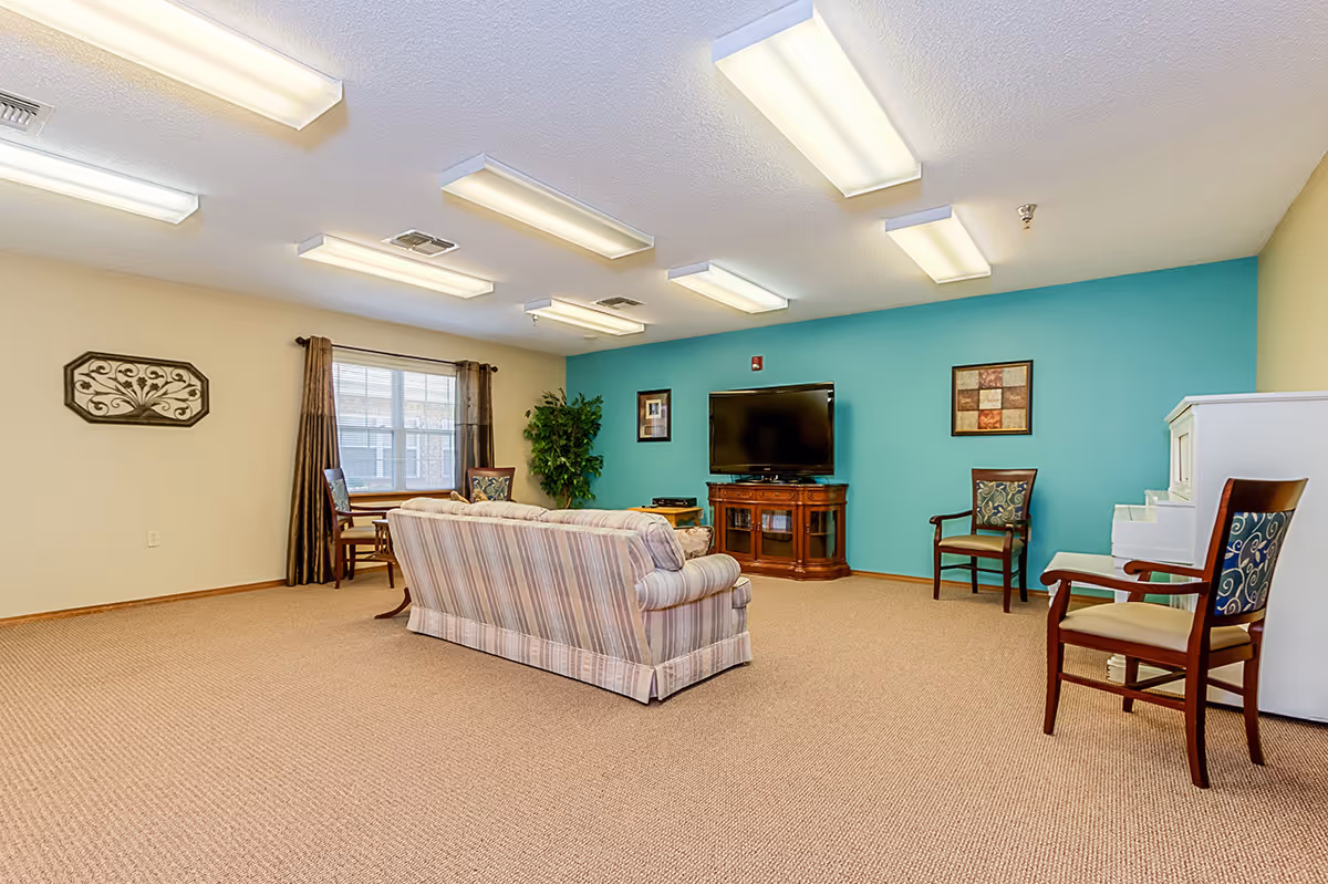 A spacious senior living common area with beige carpet and walls painted in beige and teal. The room features a striped sofa facing a flat-screen TV on a wooden stand, several wooden chairs with patterned cushions, a white piano, a window with brown curtains, and a decorative wall hanging. The ceiling has multiple fluorescent lights.