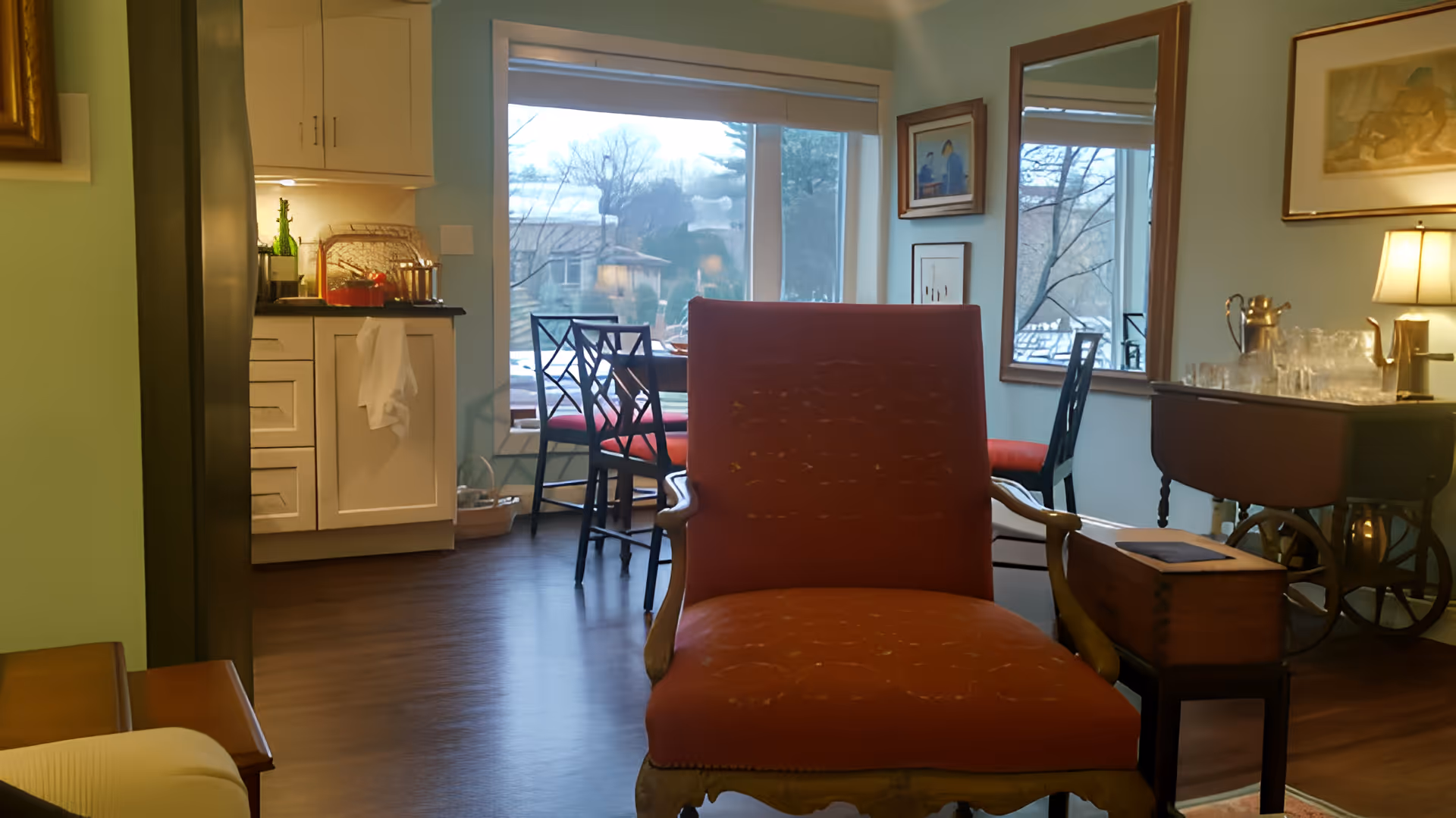 Interior view of a cozy living area with a red upholstered armchair in the foreground, a dining table with chairs near a large window showing an outdoor scene, a small kitchen area with white cabinets and a refrigerator on the left, and a sideboard with a lamp, mirror, and decorative items on the right.