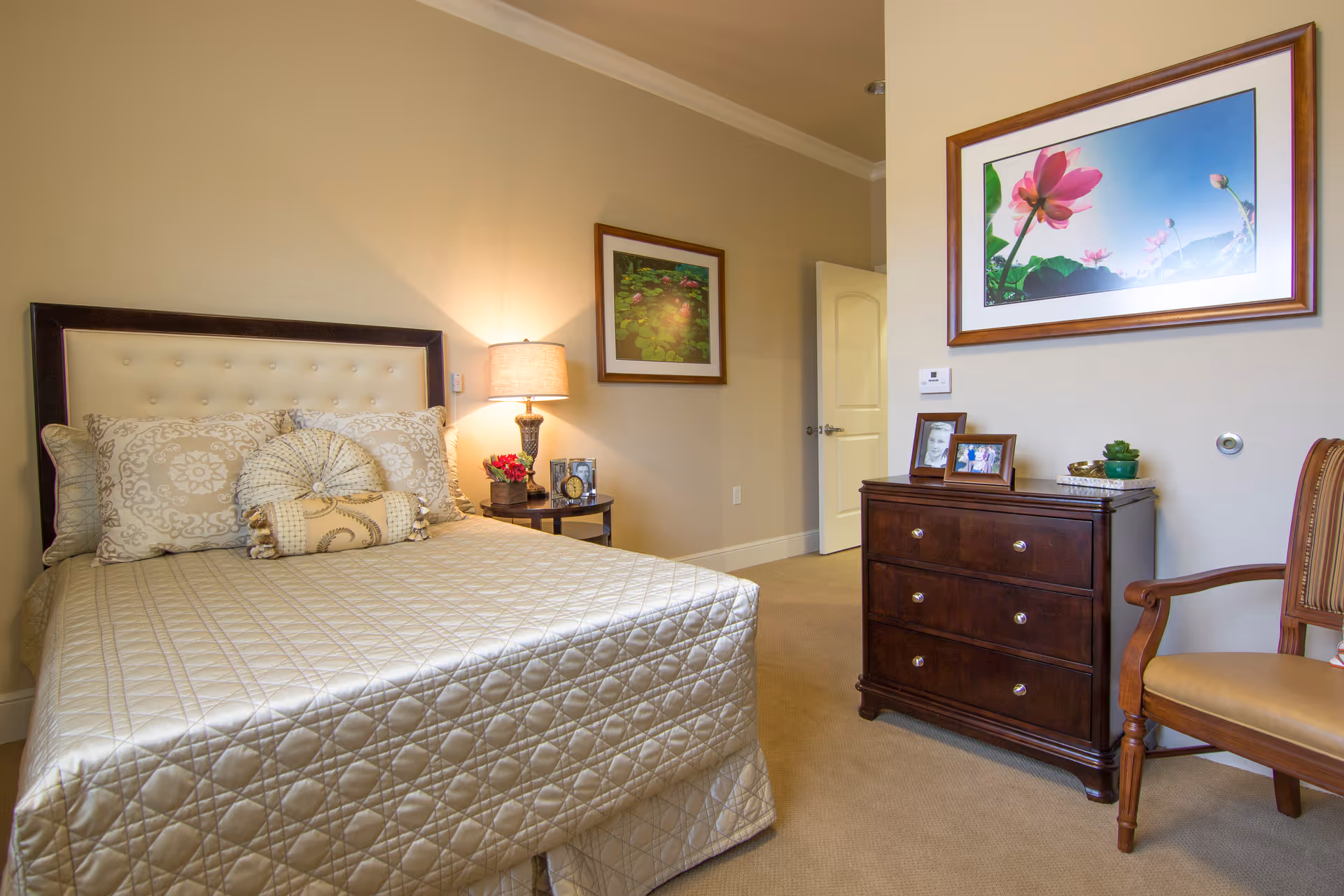 Bedroom with a bed dressed in patterned bedding, a nightstand and lamp, framed artwork, a wooden dresser, and an upholstered chair.