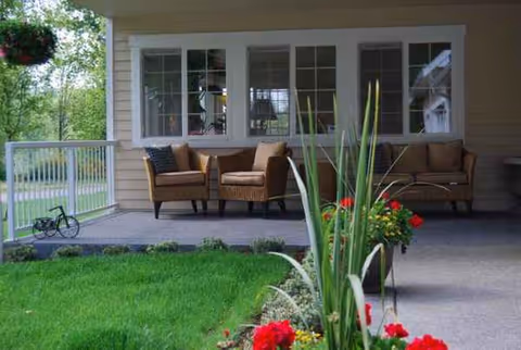 Covered porch with outdoor wicker seating, potted plants, and red flowers.