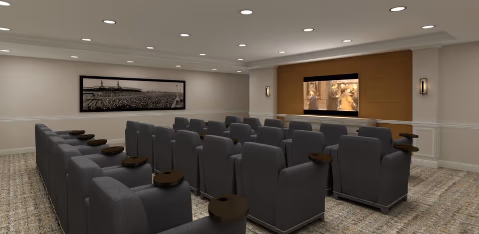 A small movie theater room with multiple rows of gray cushioned recliner chairs, each equipped with a small round wooden side table. A large flat-screen TV is mounted on a wooden accent wall at the front, displaying a scene from a movie. The room has recessed ceiling lights and a framed black and white panoramic photograph on one side wall.