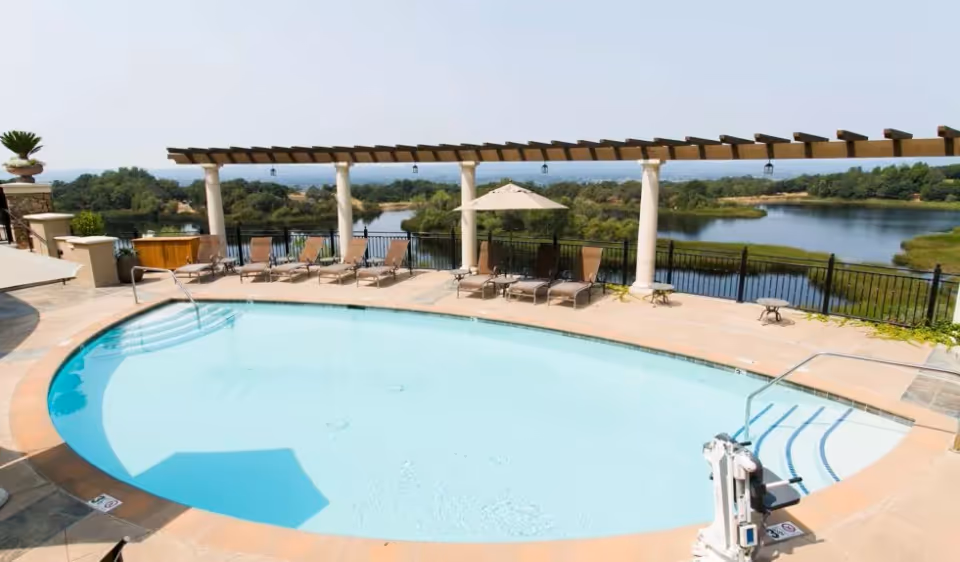Outdoor swimming pool with lounge chairs and a pergola overlooking a scenic lake and greenery.