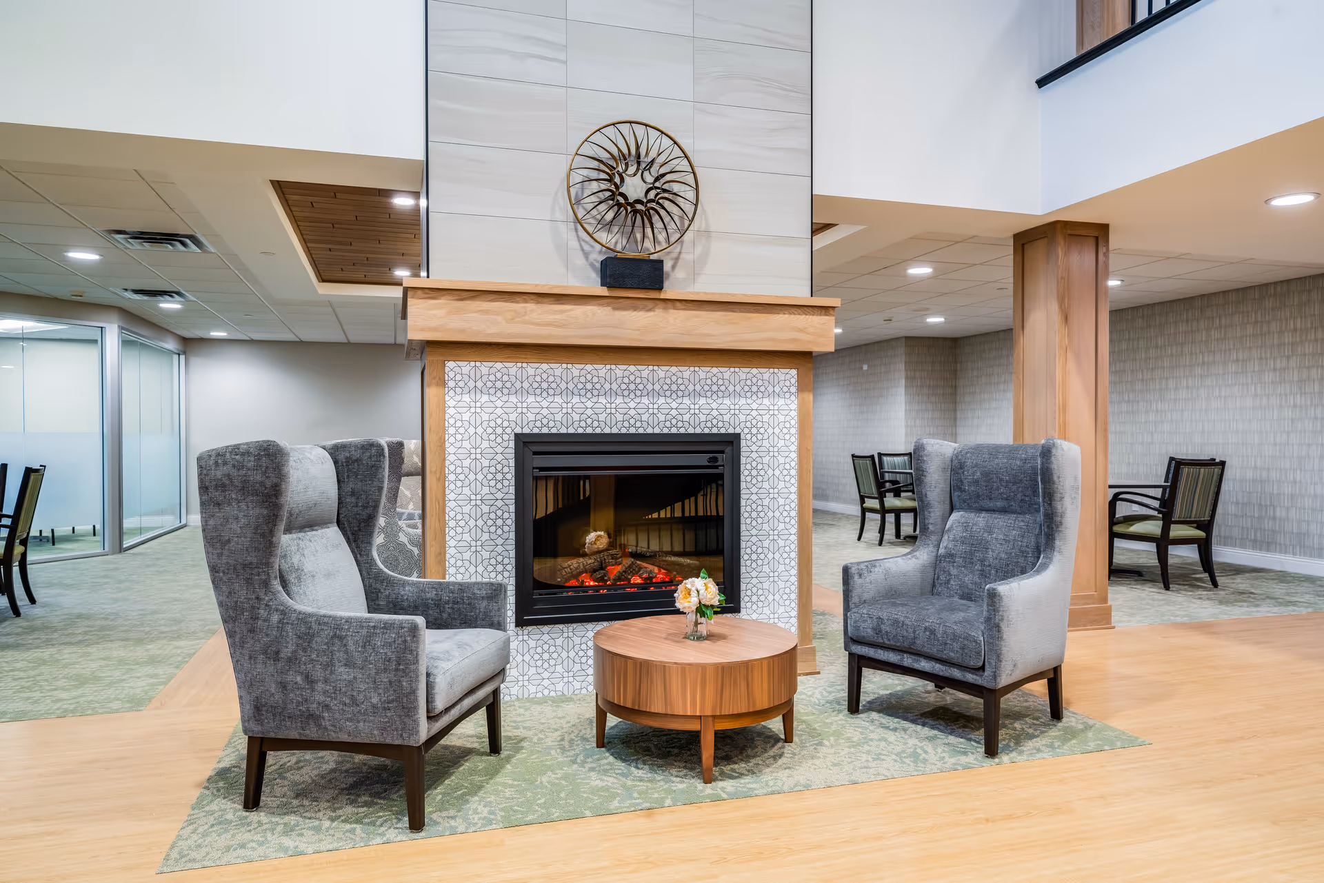 Two gray wingback chairs and a small round table face a fireplace in a modern senior living common area.