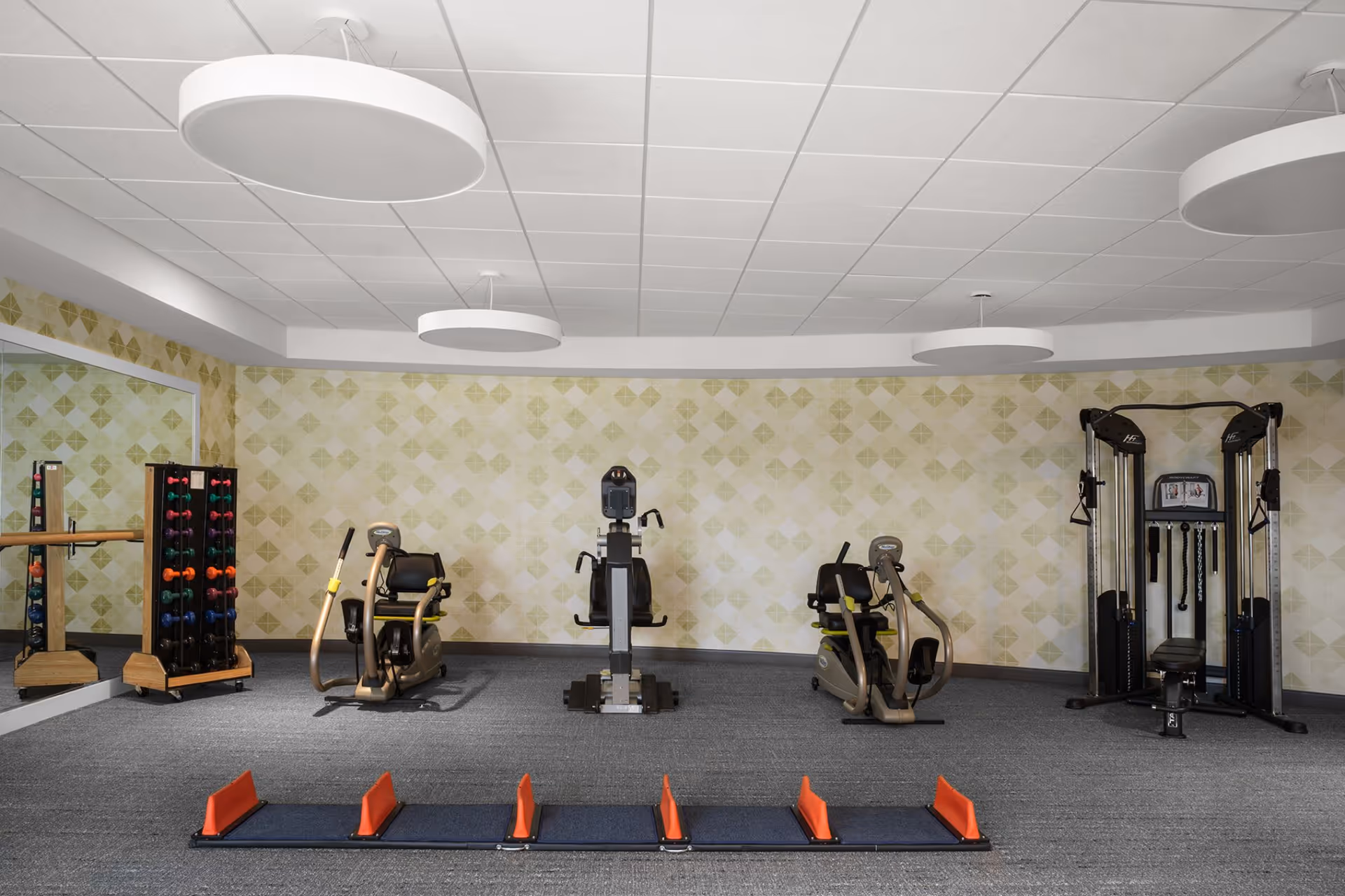 A fitness room with exercise equipment including two recumbent bikes, a multi-functional weight machine, a rack of colorful dumbbells, and a mirrored wall. The room has a patterned light green and beige wallpaper, gray carpet, and three large circular ceiling lights.