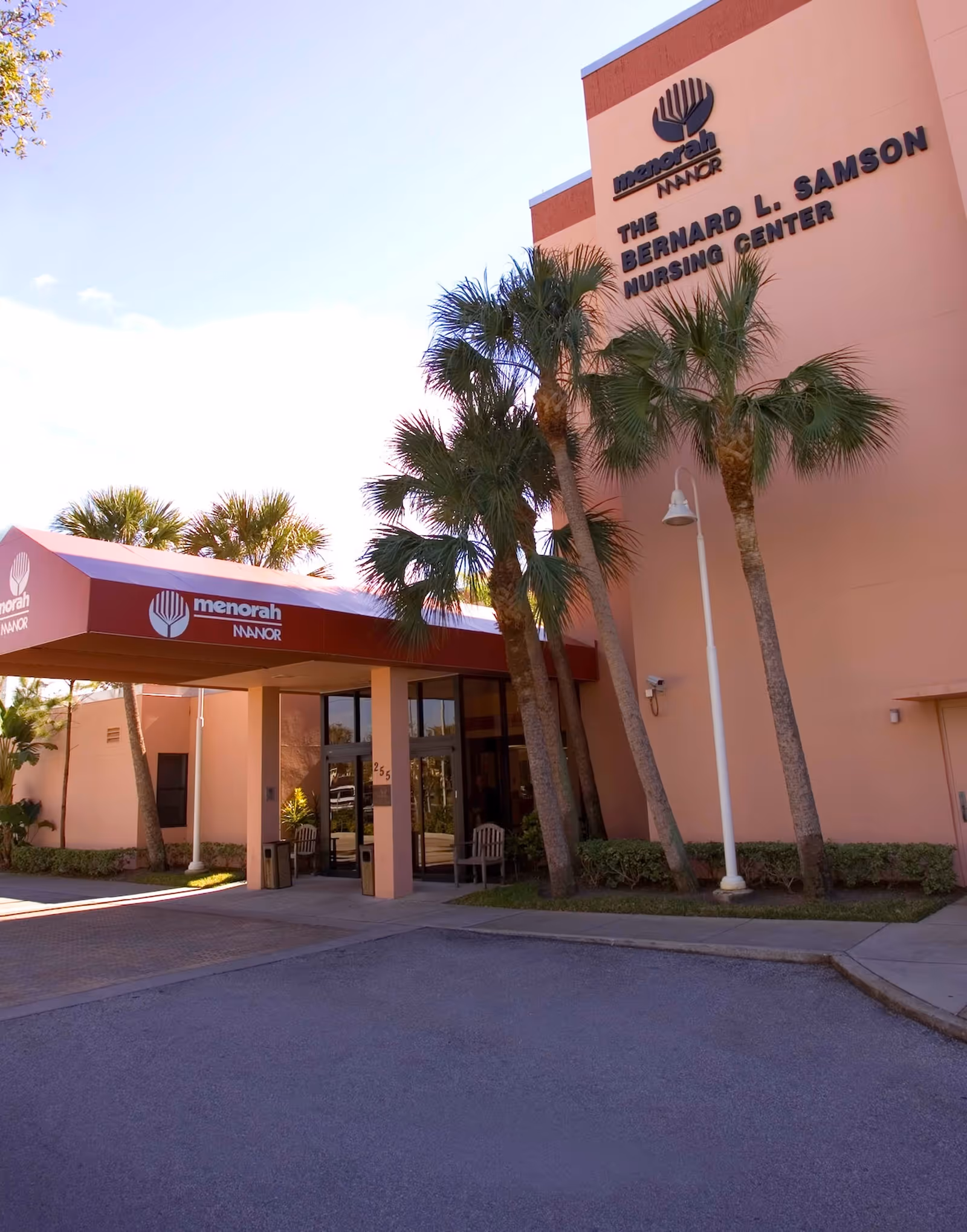 Front entrance of the Menorah Manor Bernard L. Samson Nursing Center with a covered drop-off, palm trees, and visible building signage.