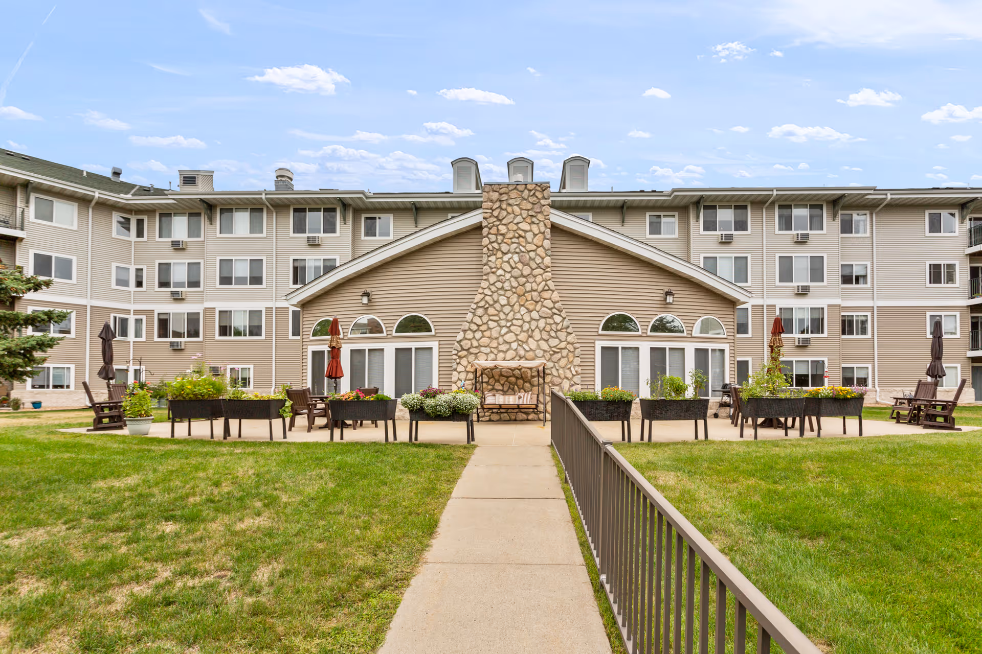 Outdoor view of Westridge Retirement Community showing a large multi-story building with beige siding and many windows. In the foreground, there is a concrete pathway leading to a patio area with a stone fireplace, outdoor seating, potted plants, and umbrellas. The sky is partly cloudy.