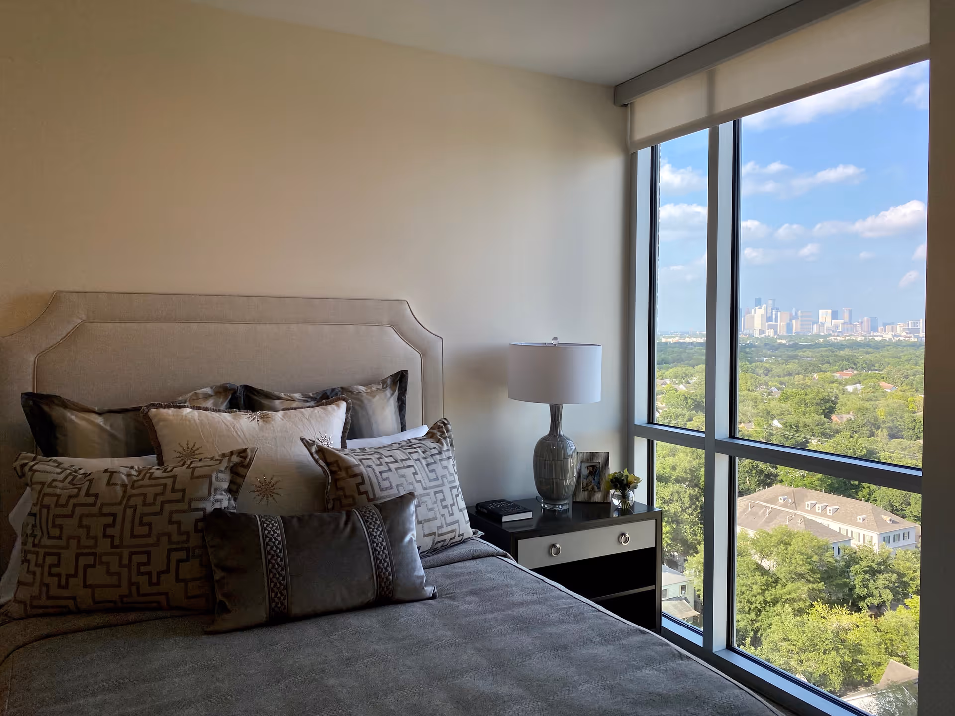 A bedroom with a neatly made bed featuring multiple decorative pillows and a beige upholstered headboard. Next to the bed is a nightstand with a table lamp, a small framed photo, and a small plant. Large floor-to-ceiling windows provide a view of a green landscape and a city skyline in the distance under a partly cloudy sky.