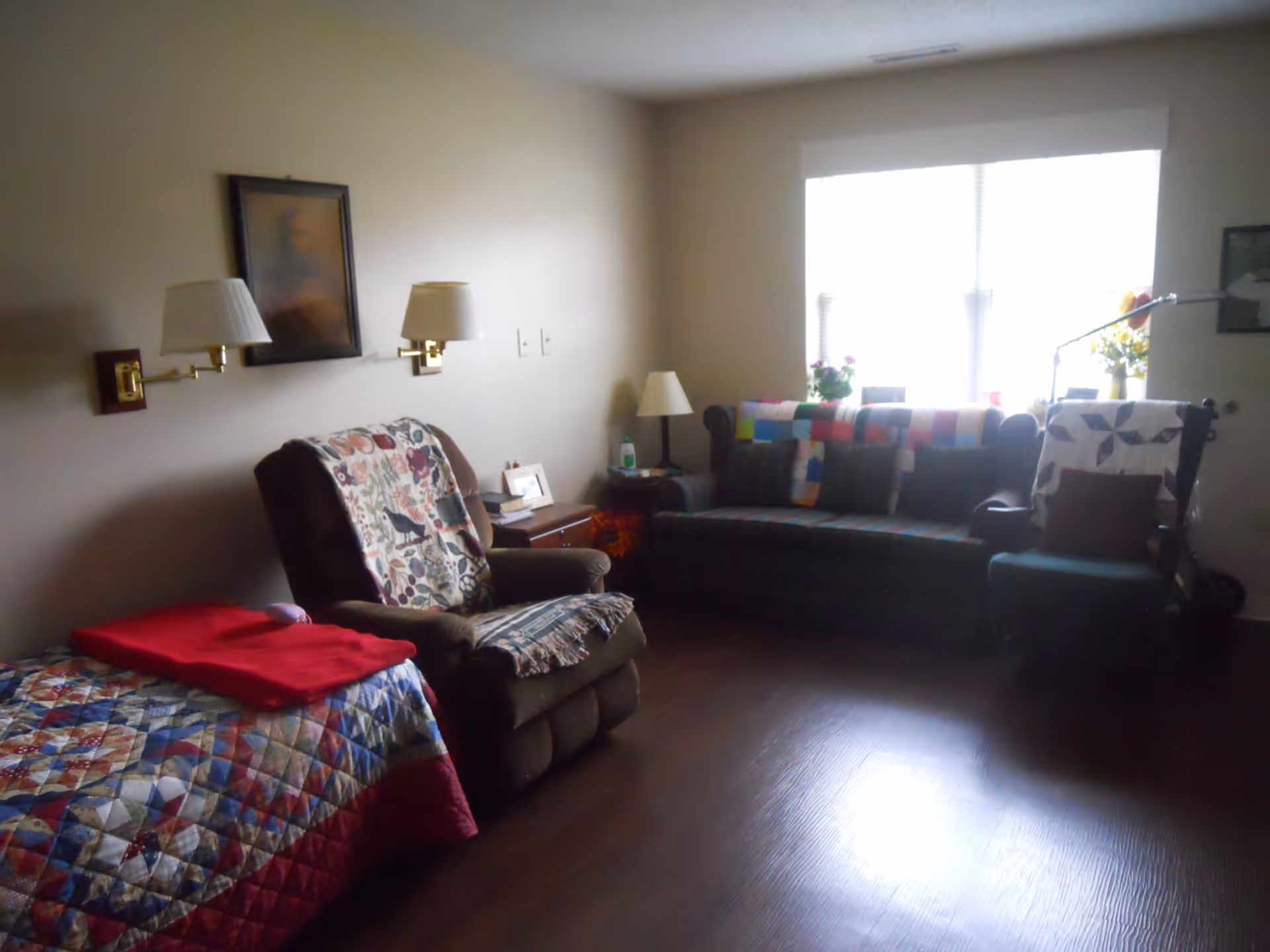 A cozy living room with a recliner chair covered with a floral quilt, a plaid sofa with colorful cushions and a crocheted blanket, a wooden side table with a lamp and framed photo, and a window letting in natural light. The floor is wooden and the walls are light-colored with two wall-mounted lamps and a framed picture.