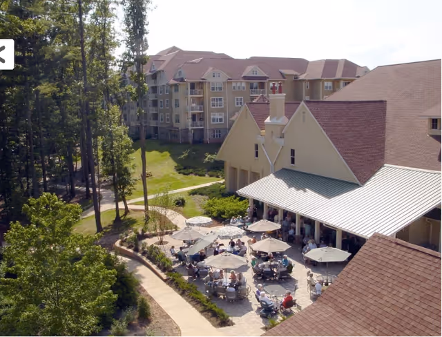 Outdoor patio area at Deerfield Episcopal Retirement Community with multiple tables and umbrellas where people are seated and socializing. The patio is adjacent to a large building with a sloped roof, surrounded by trees and landscaped pathways. In the background, there are multi-story residential buildings.