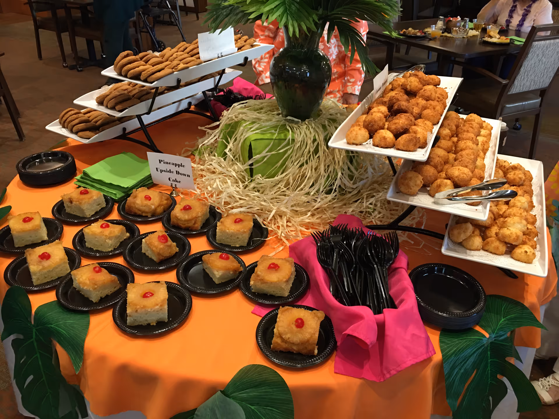 A buffet table with an orange tablecloth decorated with green leaves and a centerpiece with a green vase and straw-like material. The table displays trays of cookies, pineapple upside down cake squares topped with cherries on black plates, and several trays of golden brown fried snacks. Black plastic forks and small black plates are also arranged on the table. In the background, people are seated at dining tables.