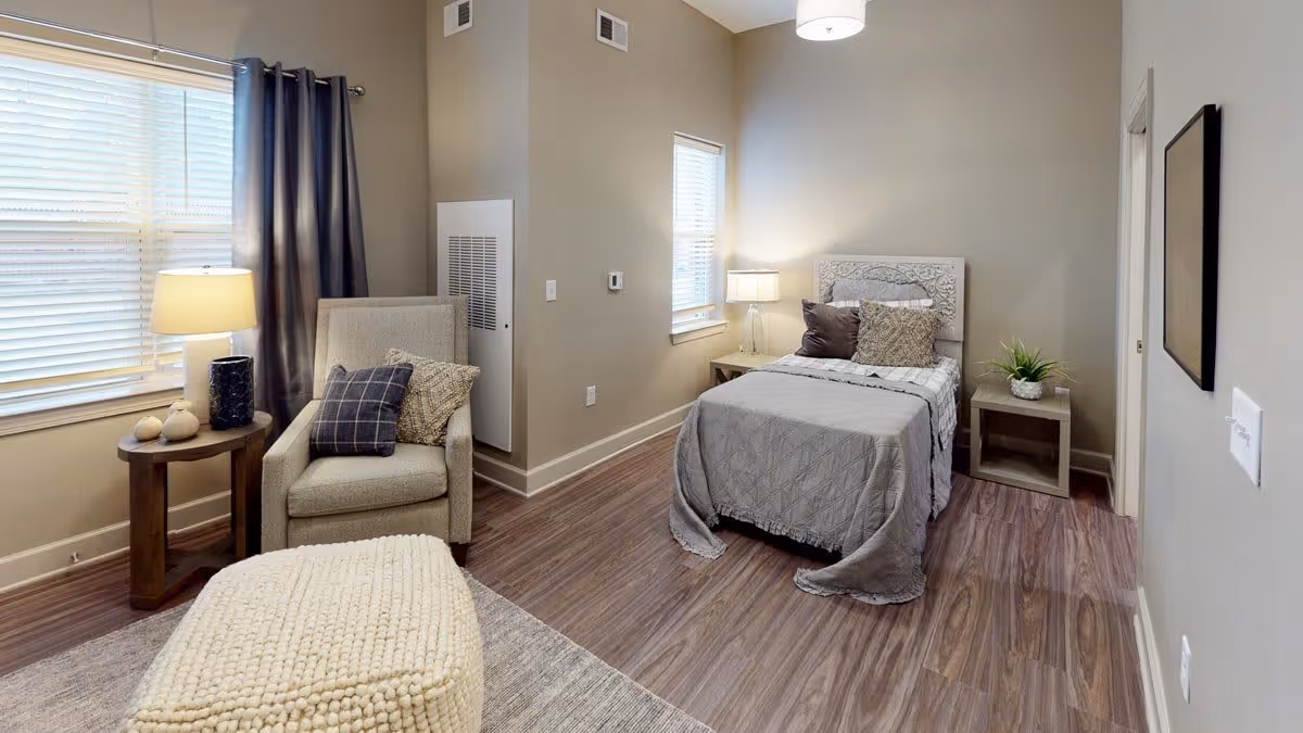 Neatly furnished bedroom with a single bed, armchair, side tables and soft lighting in neutral tones.