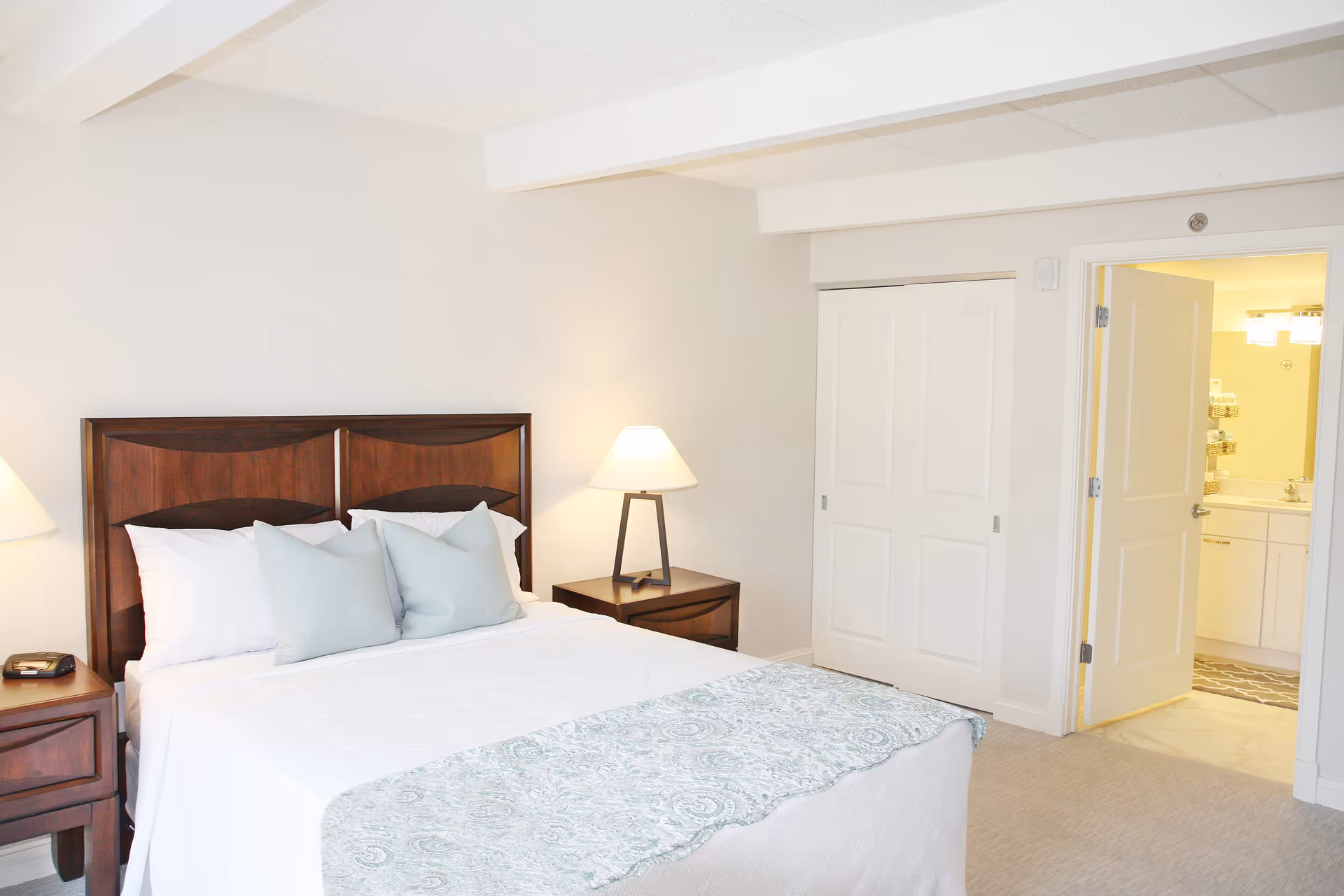 A bright bedroom with a neatly made bed featuring white linens and light blue pillows. The bed has a dark wooden headboard and matching nightstands on each side, each with a lamp. To the right, there is a closet with sliding white doors and an open door leading to a bathroom with a sink and mirror visible.