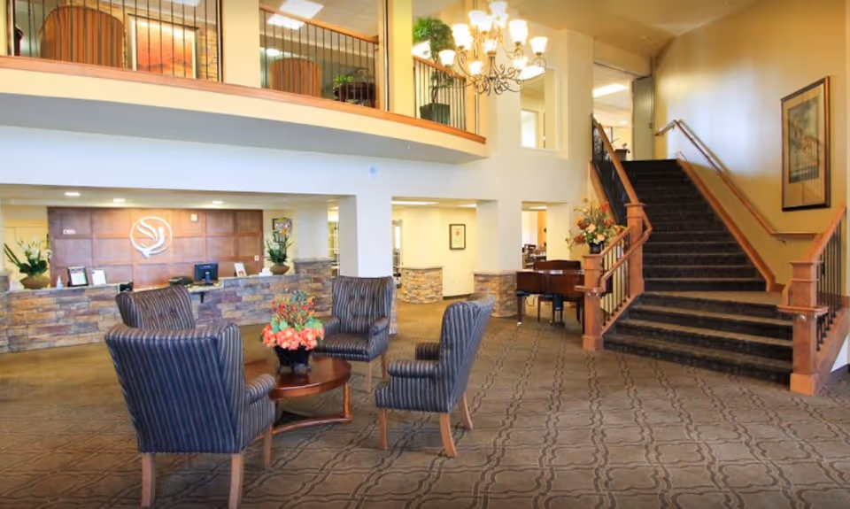 A spacious and well-lit lobby area with a seating arrangement consisting of four striped armchairs around a wooden coffee table with a floral centerpiece. Behind the seating area is a reception desk with a stone facade and a wooden panel wall featuring a logo. To the right, there is a carpeted staircase with wooden railings leading to an upper floor. The space is decorated with plants, flowers, a chandelier, and framed artwork on the walls.