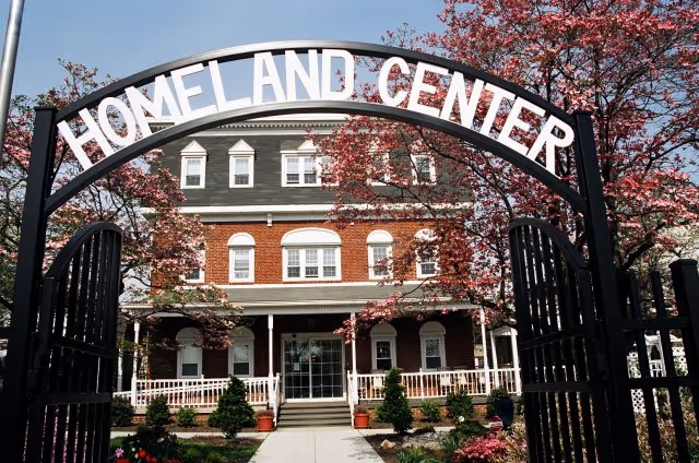 Wrought-iron entrance arch reading 'HOMELAND CENTER' leading to a brick three-story residence framed by flowering trees.