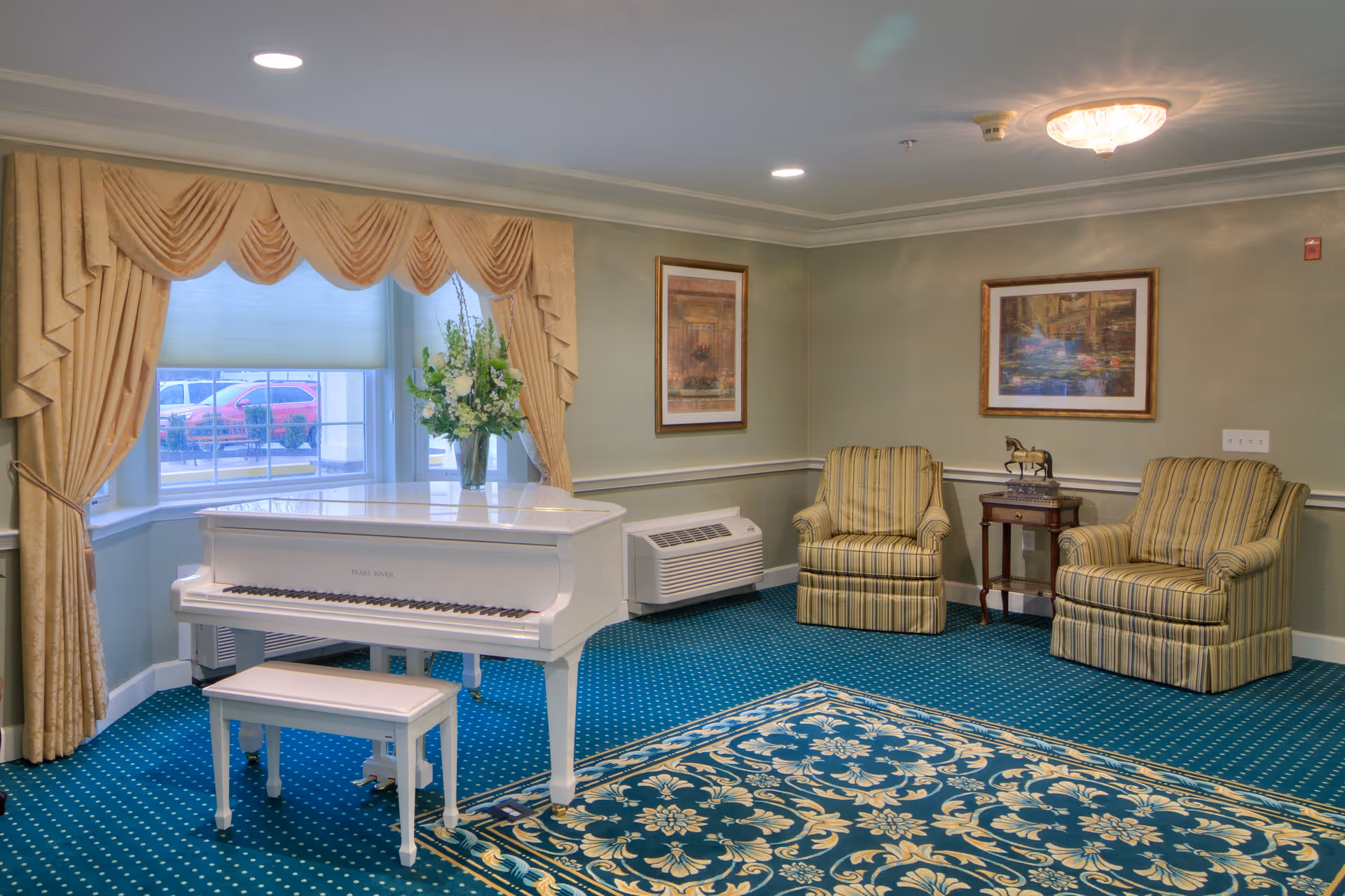 A cozy sitting area with a white grand piano and matching bench near a window with beige drapes. Two striped armchairs flank a small wooden side table with a horse figurine. The room has a blue patterned carpet and two framed paintings on the walls.