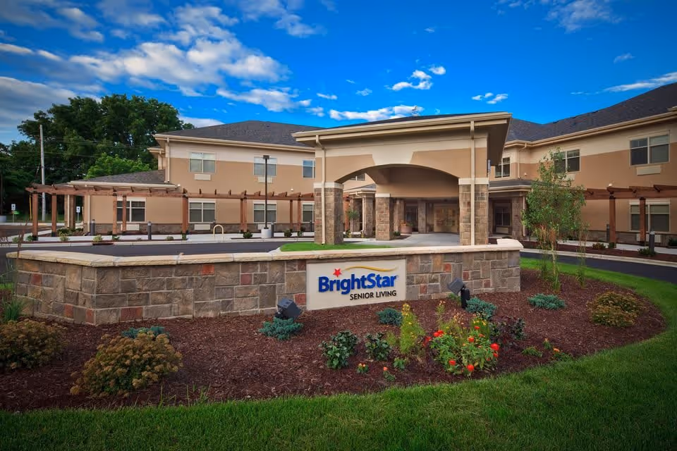 Exterior view of BrightStar Senior Living facility with a stone and brick entrance sign surrounded by landscaping, a covered entryway, and a two-story building under a partly cloudy blue sky.