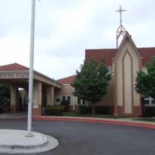 Exterior view of Bishop Spencer Place facility showing a building with a peaked roof and a cross on top, surrounded by trees and a driveway with a flagpole in the foreground.
