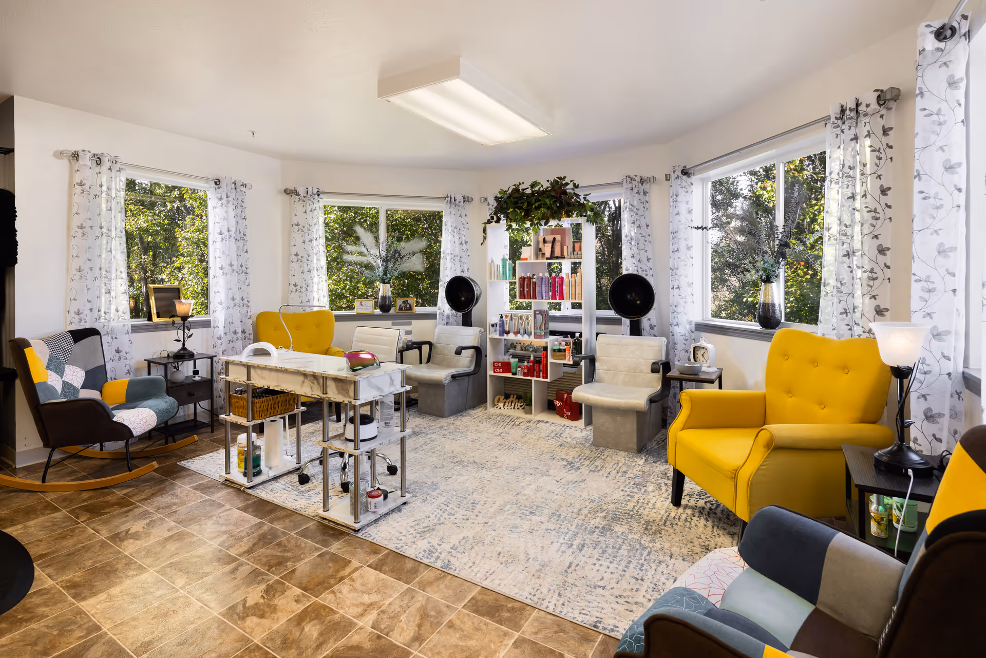A bright and cozy salon area in a senior living community with large windows covered by sheer white curtains with a leaf pattern. The room features a mix of colorful armchairs including yellow and patchwork designs, a white rolling cart with beauty supplies, two hair dryer chairs, a white shelving unit stocked with hair care products, and decorative plants on the windowsills.