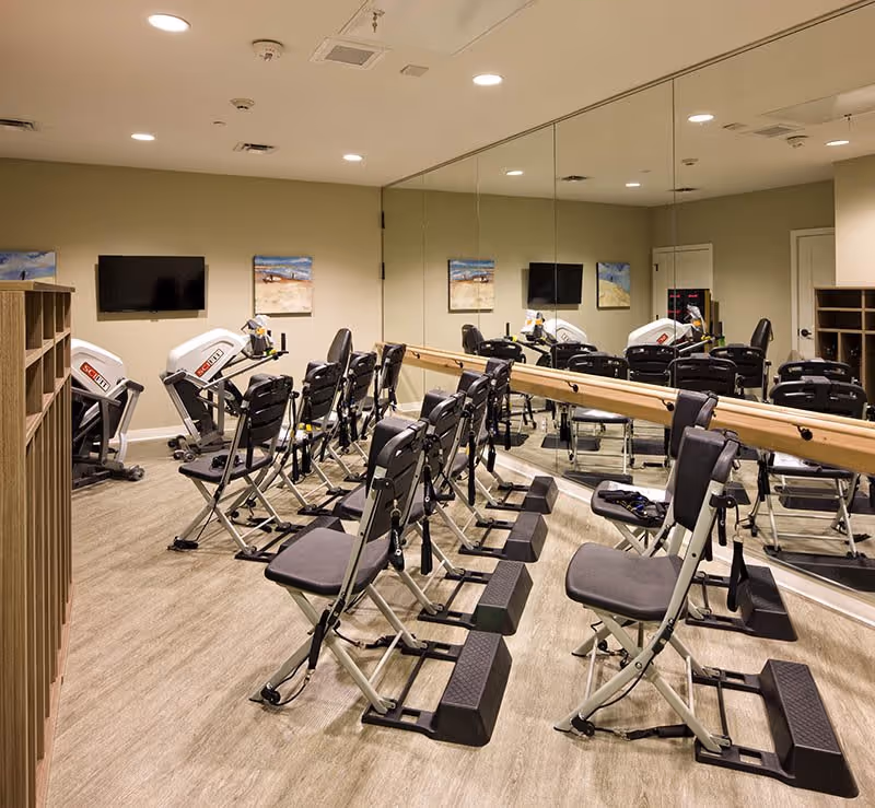 Exercise room with multiple seated exercise machines arranged in rows facing a wall-mounted TV and two framed pictures. The room has a large mirror covering one wall and a wooden ballet barre attached to the mirrored wall. The floor is wood-style, and the ceiling has recessed lighting.