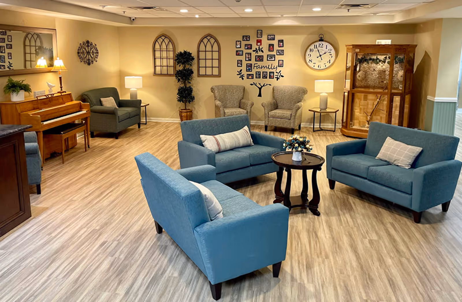 A cozy living room area with three blue upholstered loveseats arranged around a small round wooden table with a flower vase. The room has light wood flooring and beige walls decorated with a large clock, family photo collage, two arched window-like wall decorations, and a tall potted plant. There are two beige armchairs with a side table and lamp, a piano with a lamp and plant on top, and a wooden display cabinet in the corner.