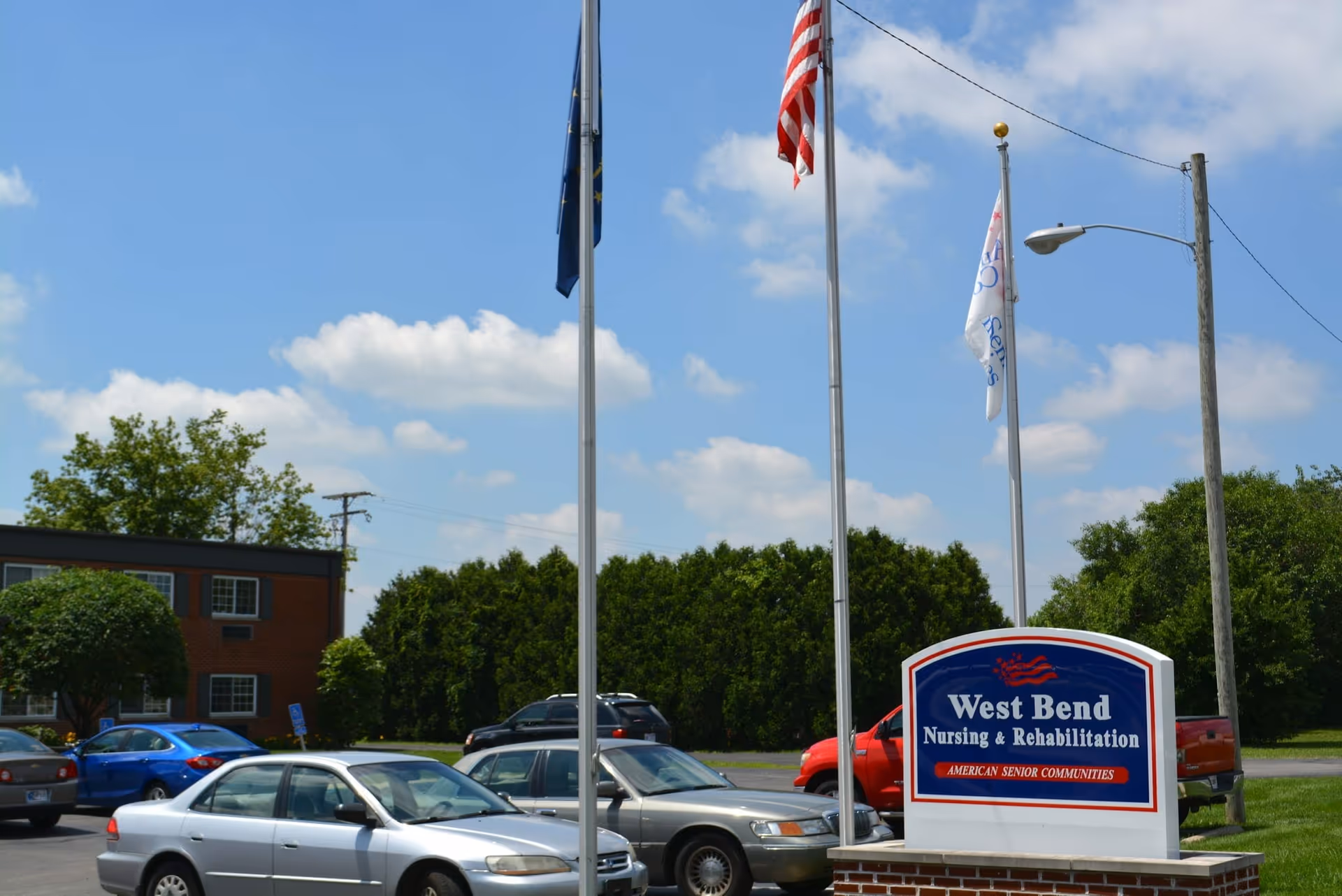 Outdoor view of the parking lot at West Bend Nursing & Rehabilitation facility with several parked cars, three flagpoles with flags, and a blue and white sign displaying the facility's name and affiliation with American Senior Communities under a partly cloudy sky.