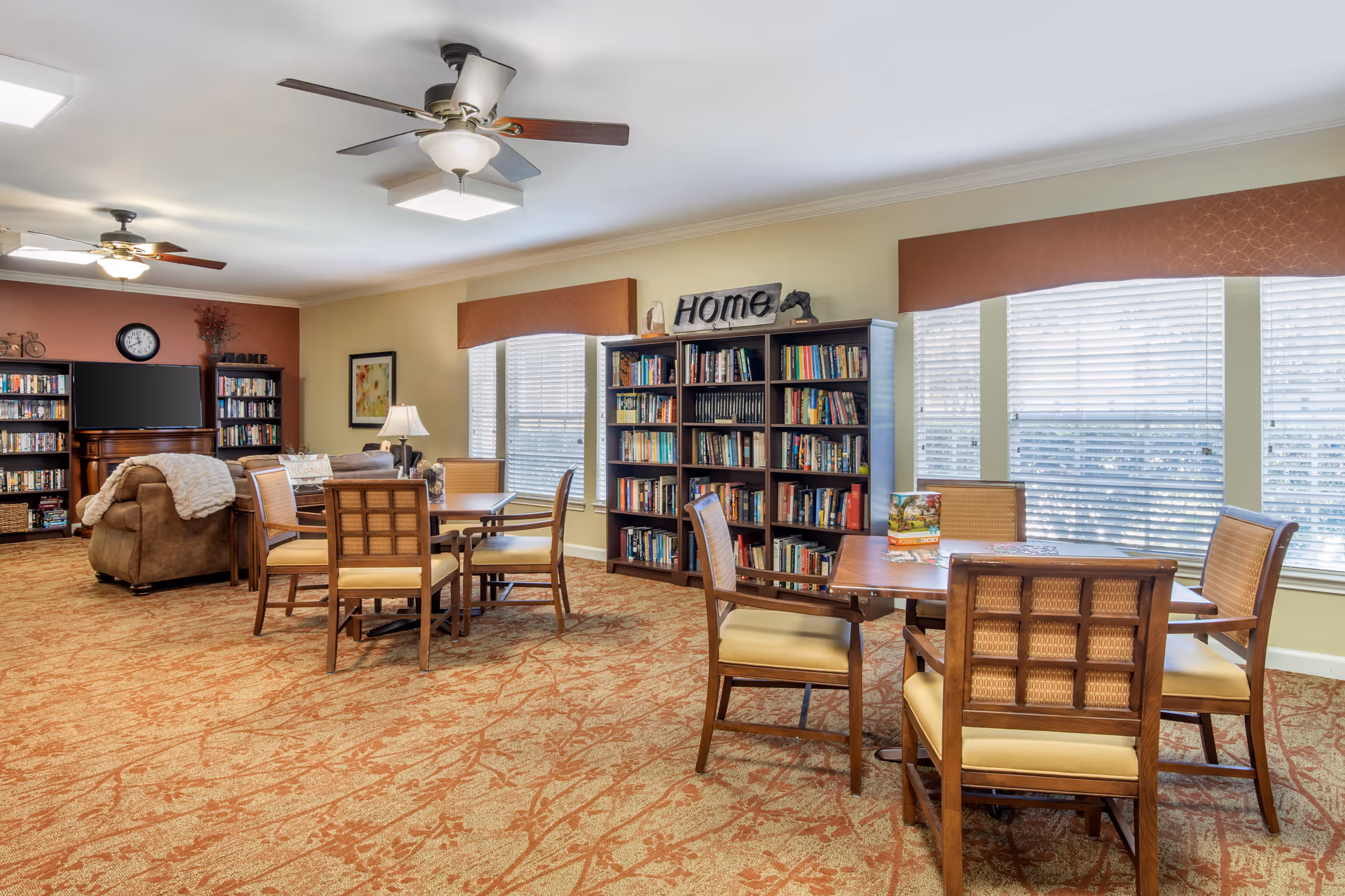A cozy common area in a senior living facility with carpeted floor, two ceiling fans, and large windows with blinds. The room features multiple wooden tables with cushioned chairs, bookshelves filled with books, a flat-screen TV mounted on a wooden stand, and decorative items including a sign that says 'Home'.