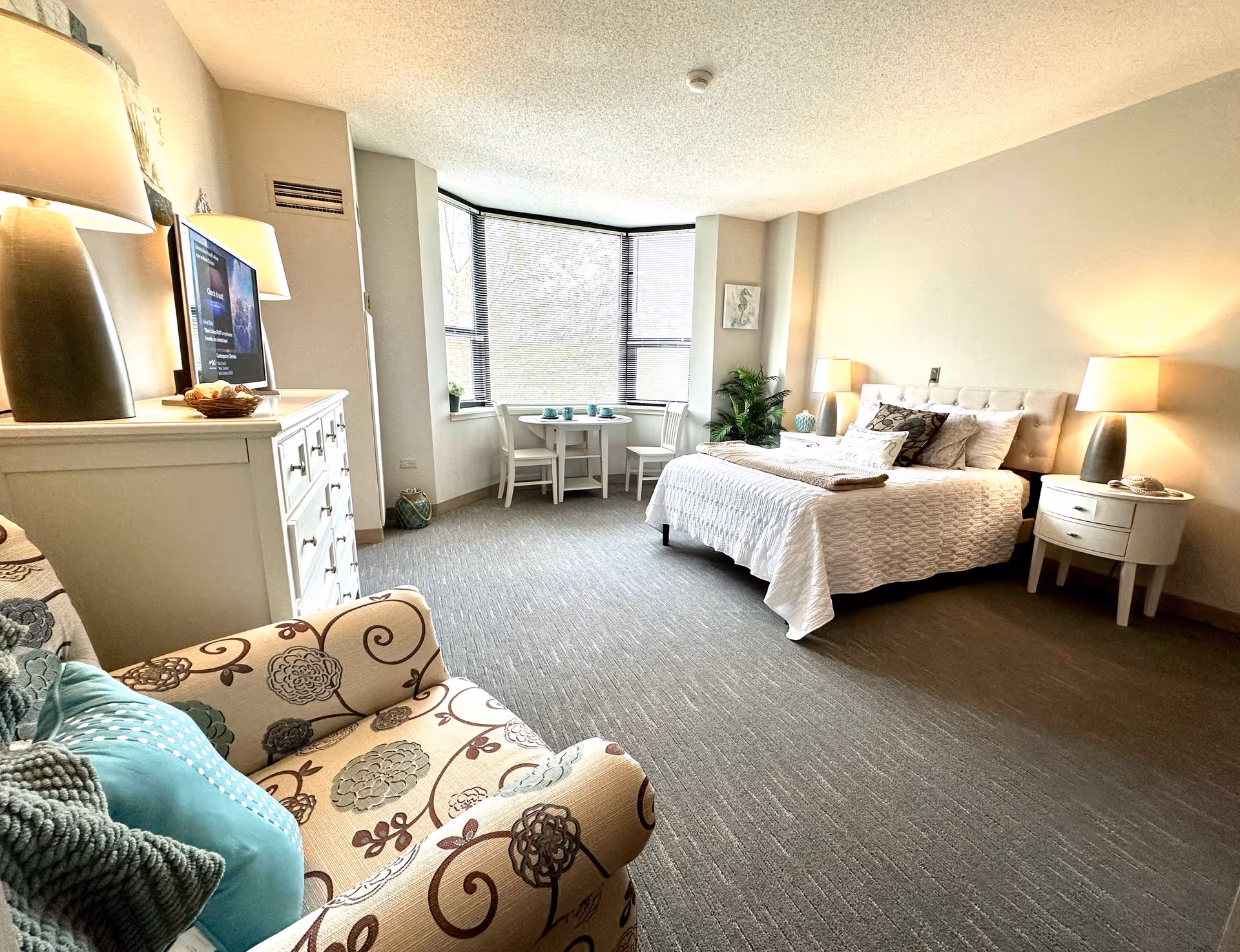 A bright and spacious bedroom in a retirement facility featuring a large bed with white bedding and multiple pillows, two bedside tables with lamps, a patterned armchair with blue and gray cushions, a white dresser with a TV on top, and a small round table with two chairs near a large window with blinds.