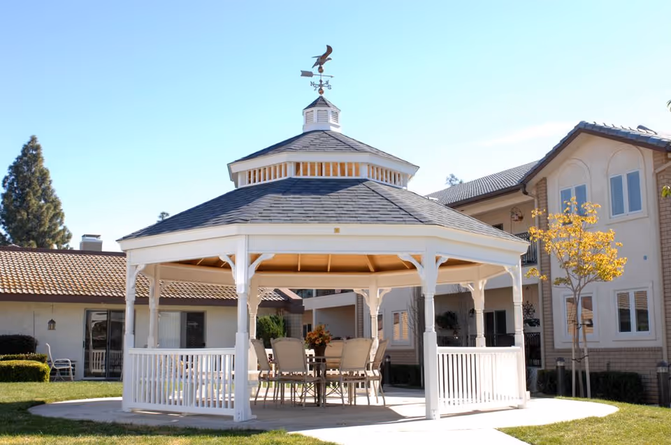 A white gazebo with a weather vane on top situated on a grassy area in front of a residential building. Inside the gazebo, there is a table with several chairs arranged around it. The building in the background has beige walls, multiple windows, and a tiled roof. A small tree with yellow leaves is visible near the gazebo.
