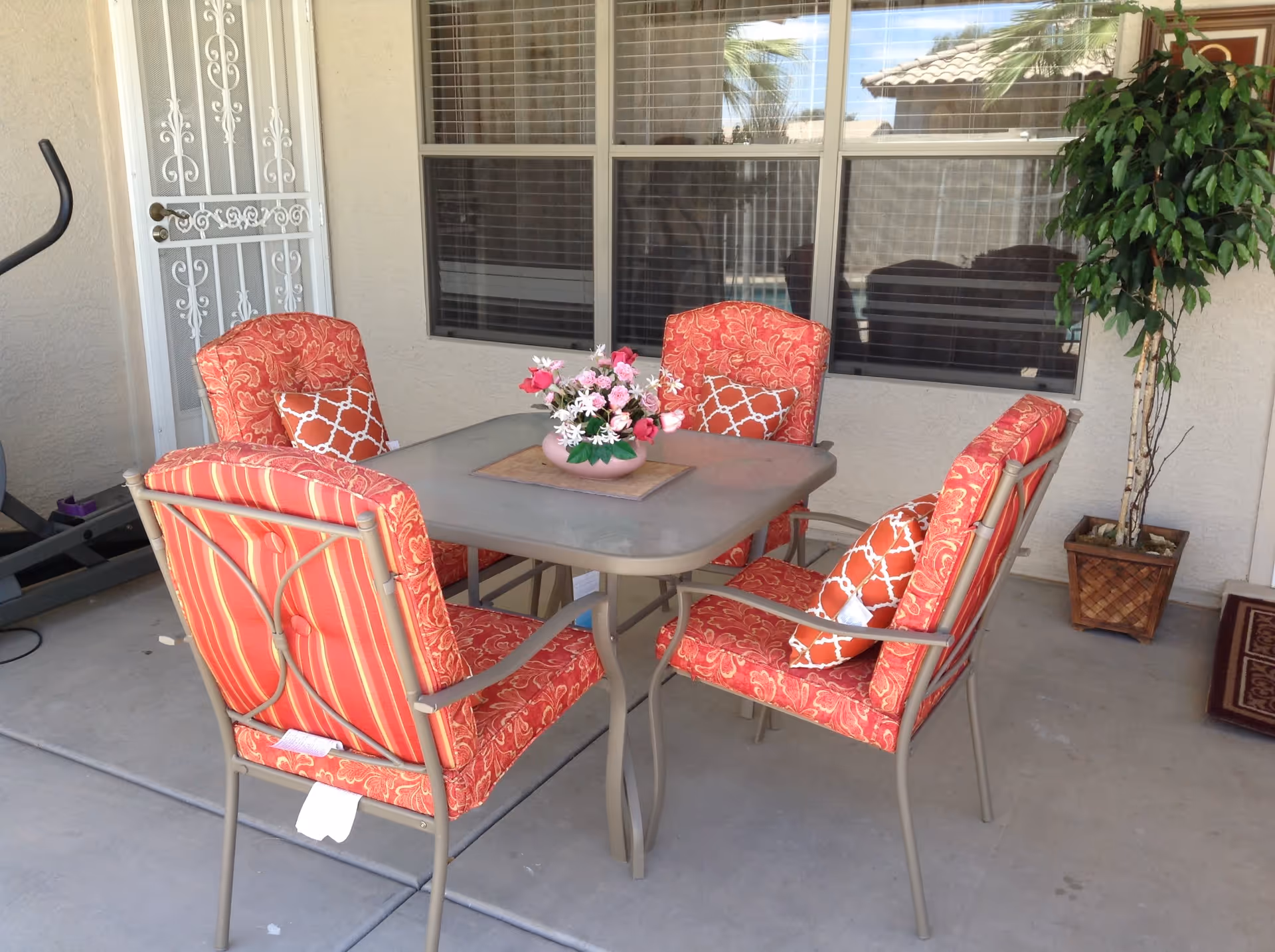 Covered outdoor patio with a square table surrounded by four red patterned cushioned chairs, a floral centerpiece, and a potted plant.