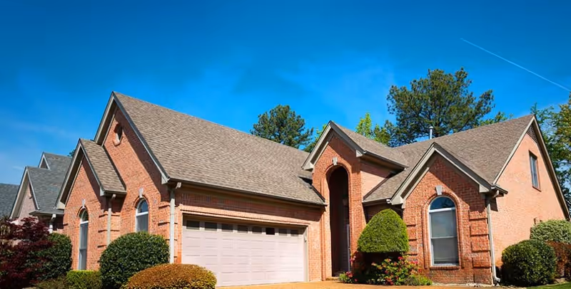 Brick single-story house with an attached garage, manicured shrubs, and a bright blue sky.